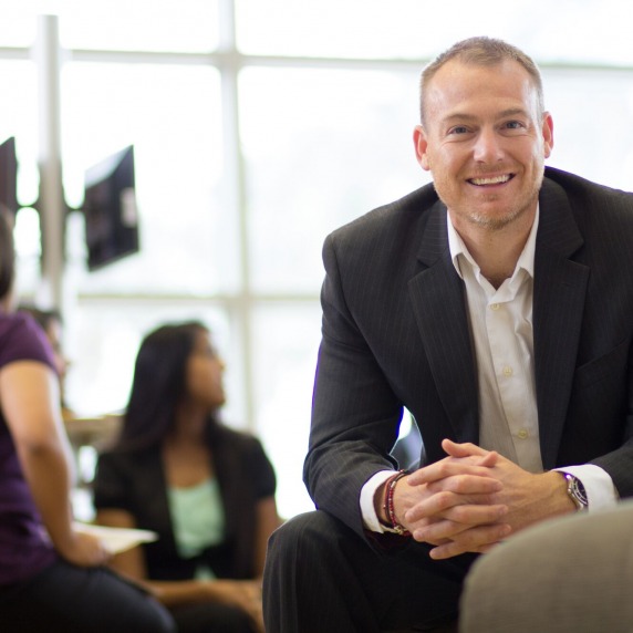Smiling white adult male wearing a business suit sitting in a lounge leaning forward with students in the background