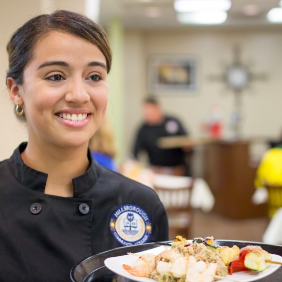 Smiling female hispanic student wearing a black chef's jacket, holding a tray of food in the Gourmet Room restaurant