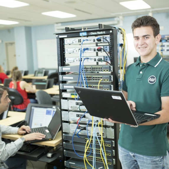 Smiling white male student holding a laptop standing next to a Cisco server rack in a classroom with other students