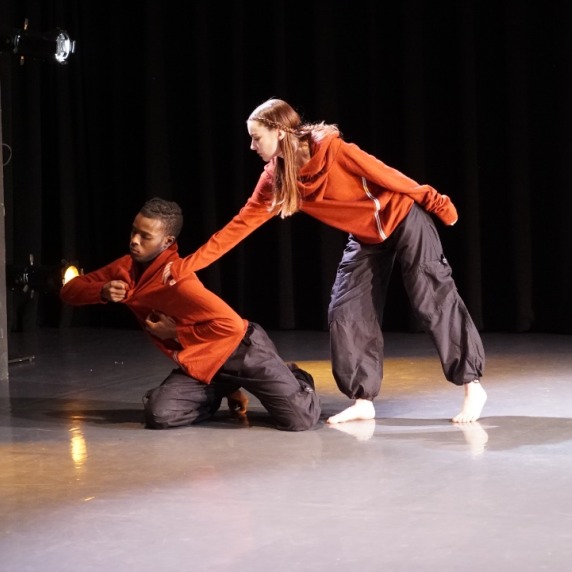 Two dancers in red sweatshirts and black sweatpants, one on his knees, the other reaching down to him.