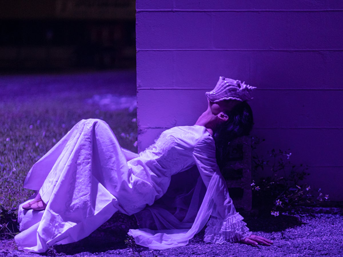 Photo of woman in long white dress lying in front of block wall with conch shell covering face