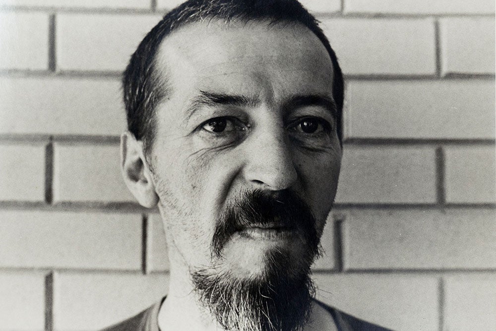 Black and white close-up portrait of a man with a beard, against a brick wall background.