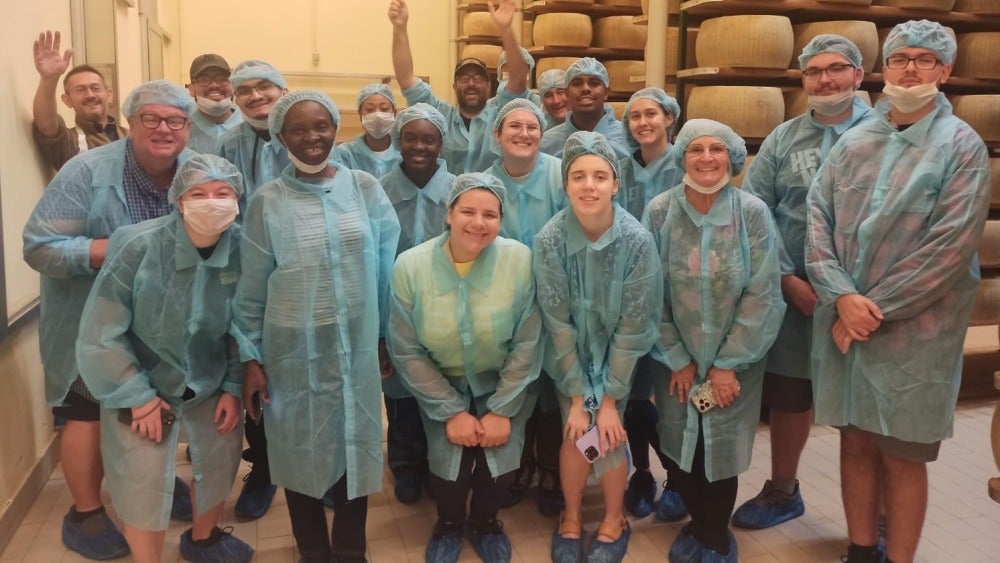 A group of people dressed in blue scrubs in front of stacks of wheels of cheese