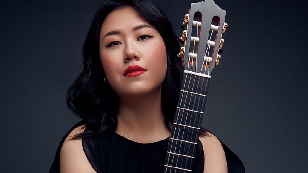 A person with black hair and red lipstick holding a classical guitar vertically.