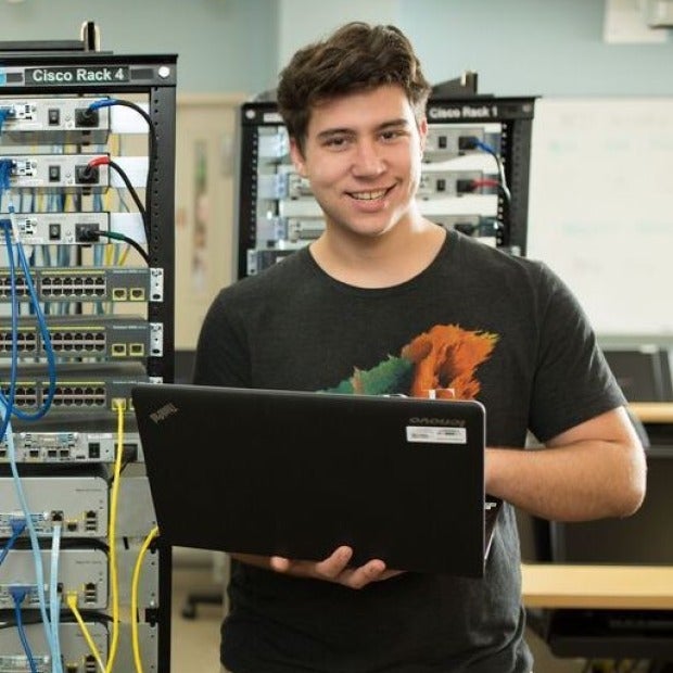 male student holding laptop next to computer servers.