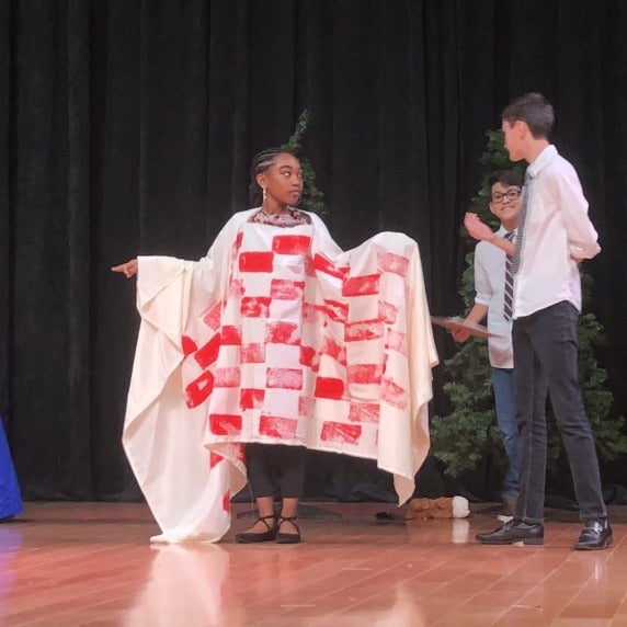 A woman on stage draped in red and black cloth, watched by two people in ties and a woman in a blue dress and green scarf