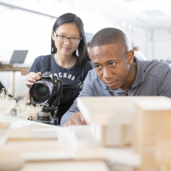 A young asian student holding a Canon camera on a tripod, next to a black male student who's looking at architectural models