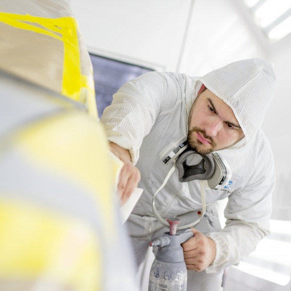Auto Collision student wearing white coverall with half-mask respirator hanging from neck while resurfacing a mask taped car