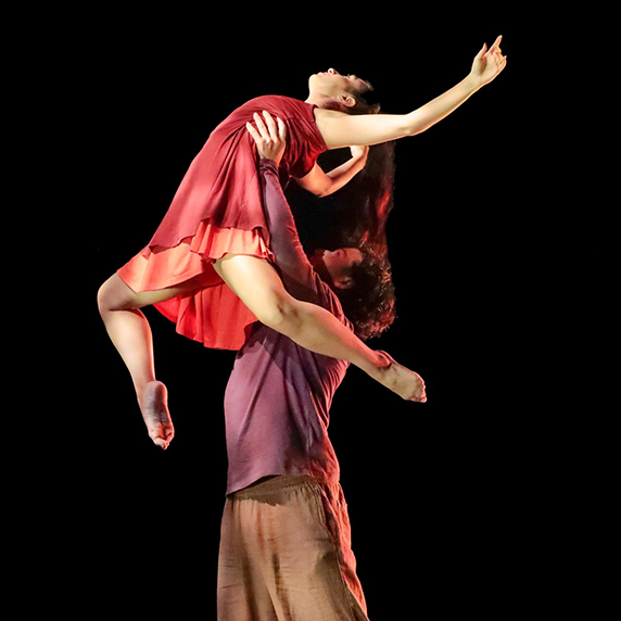 A male dancer lifts a female dancer in a red dress against a black background during a performance.
