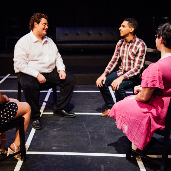 Four people seated in a circle on a stage with a dark floor and white lines.