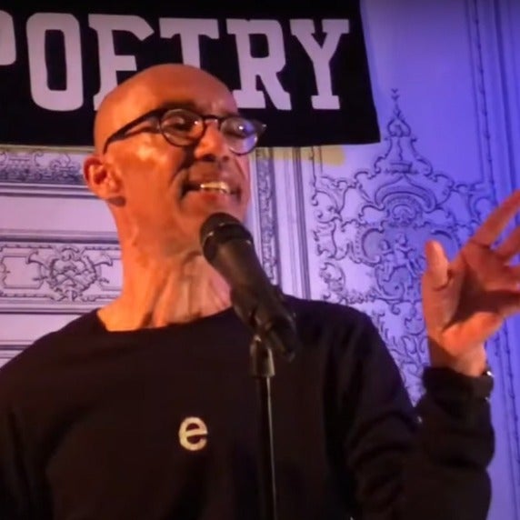 A man speaking into a microphone with a "POETRY" banner in the background.