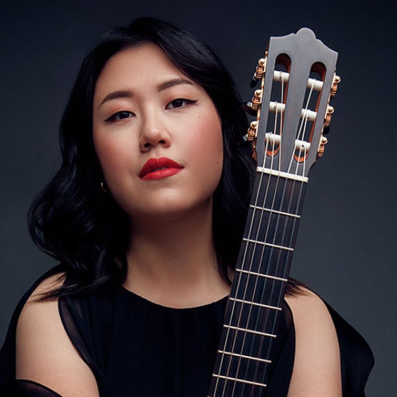 A person with black hair and red lipstick holding a classical guitar vertically.