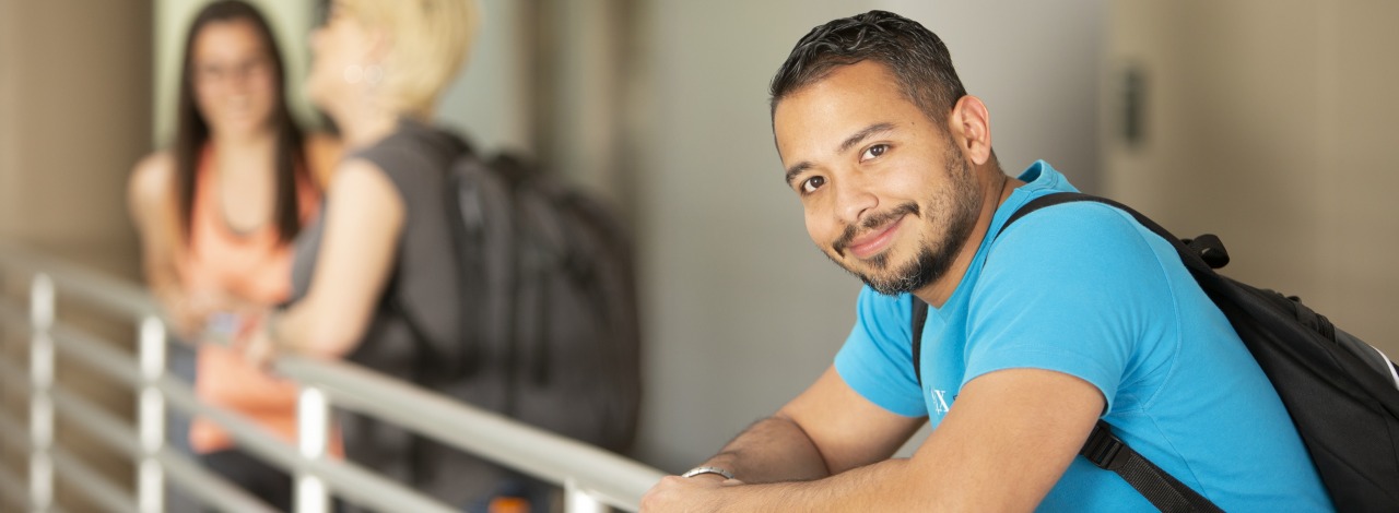 Hispanic male student smiling, leaning on a balcony rail with students in the background