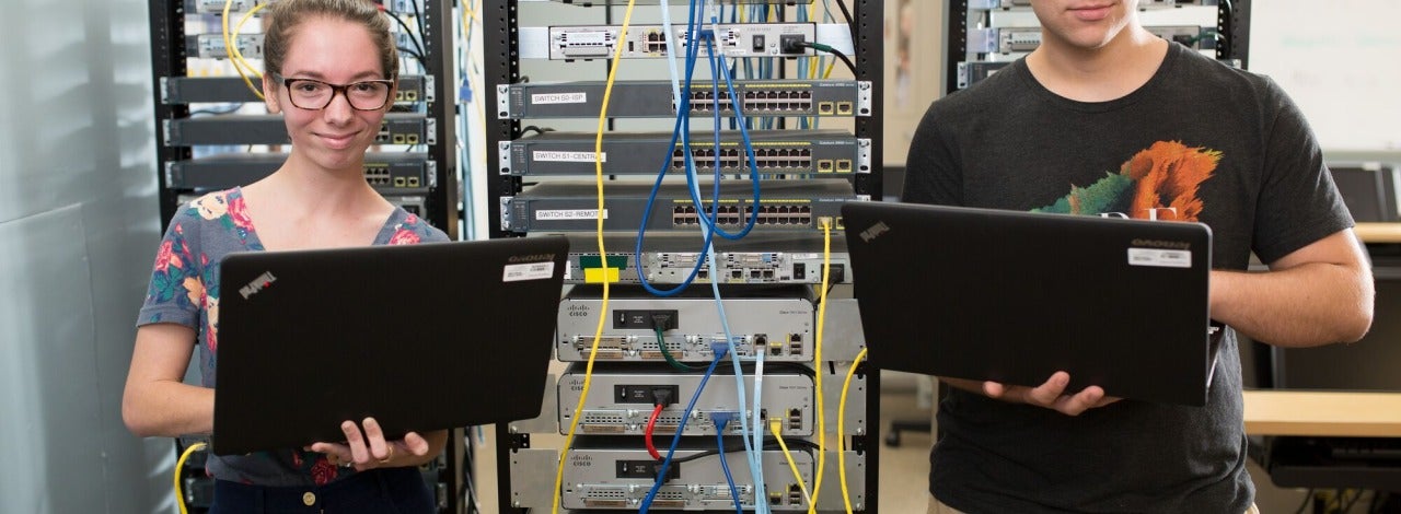 Two students  with laptops standing in front of a server rack