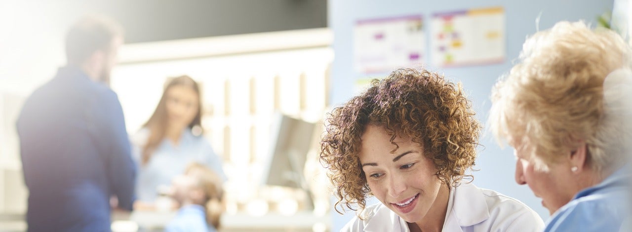 A female pharmacist sits with a senior female patient in the pharmacist consultation area and discusses her prescription and choice of medication looking on a digital tablet. In the background a father and daughter stand at the dispensing counter and are served by a female pharmacy assistant .