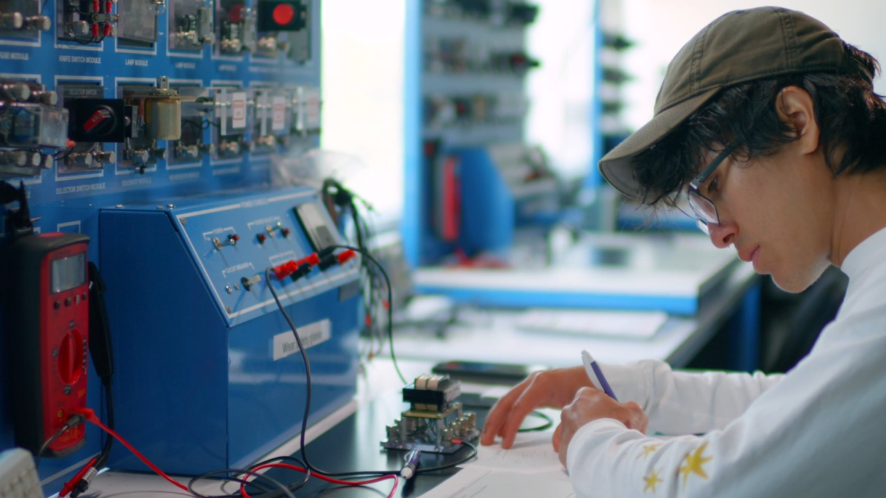 A person writing at a table with electronic equipment and a blue control panel in a laboratory.