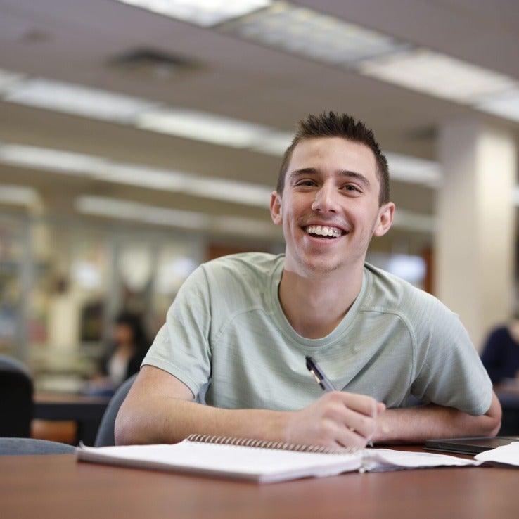 A student sitting at a table in a campus library directly smiling at the camera. 